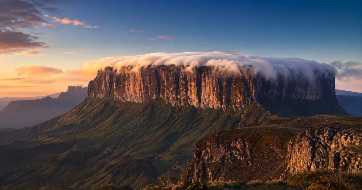 A long flat-topped massif above a coastal city at dusk, cloud spilling over its level rim